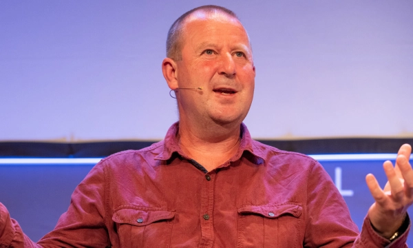 A middle-aged man with short hair and a microphone on his ear speaks passionately while gesturing with his hands on stage.