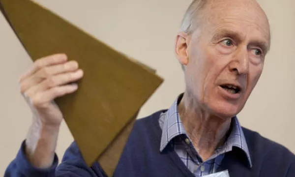 A man with gray hair speaks passionately while holding up a triangular piece of material, possibly demonstrating a concept or idea.