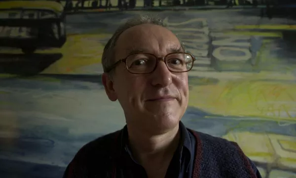 A smiling man with glasses stands in front of a colorful, abstract mural depicting a street scene with vehicles and the phrase "No Stopping."