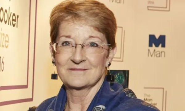 A woman with short hair and glasses smiles at an event, wearing a blue scarf, with a backdrop featuring the Man Booker Prize logo.