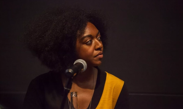 A woman with natural curly hair sits attentively at a panel discussion, microphone in front of her, wearing a black and yellow top.
