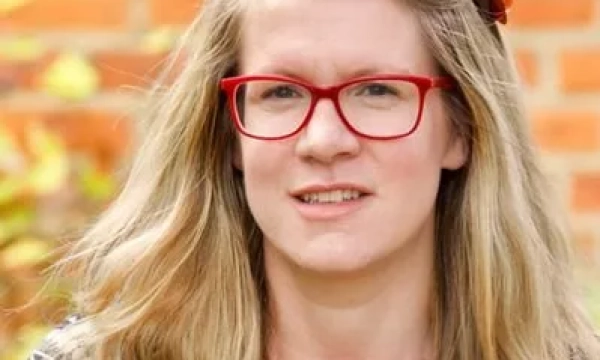 A woman with long blonde hair and red glasses, wearing a black and white patterned sweater and a colorful necklace, poses outdoors against a brick wall.