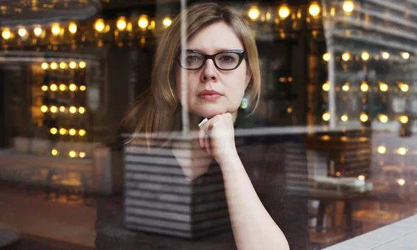 A thoughtful individual with glasses rests her chin on her hand, gazing reflectively through a glass window illuminated by warm lights.