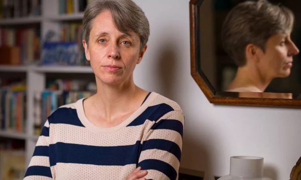 A woman with short gray hair stands with crossed arms, looking directly at the camera, with a bookshelf and a mirror reflecting her profile in the background.
