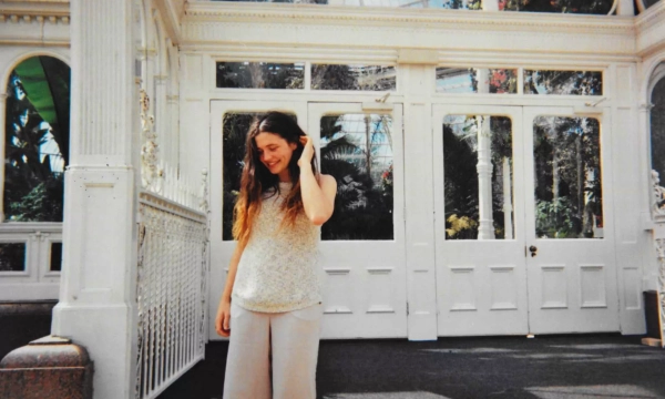 A young woman with long hair stands outside a white, ornate greenhouse, wearing a light sweater and wide-leg pants, smiling softly.