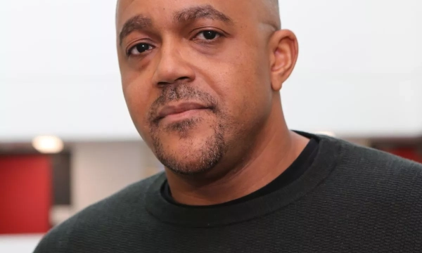 A close-up portrait of a man with short hair and a goatee, wearing a black sweater, against a modern, minimalist background.