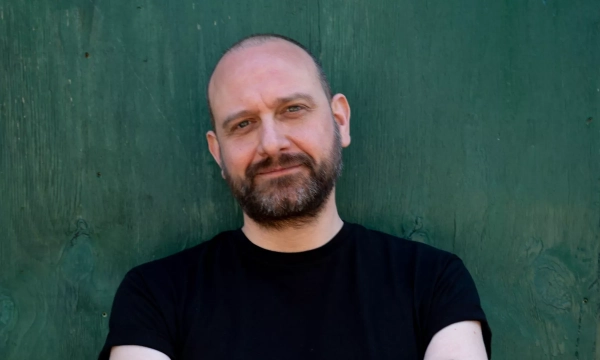 A man with a beard and bald head, wearing a black t-shirt, poses casually in front of a green wooden background.