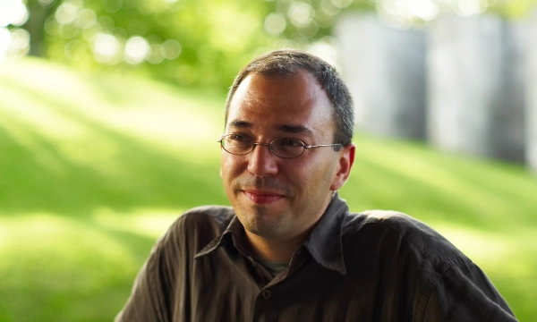 A man with glasses and short hair smiles while seated outdoors, with a blurred green background.
