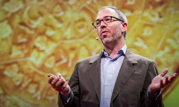 A speaker presenting on stage, gesturing with one hand while holding a microphone, with a colorful abstract background behind him.