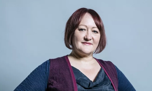A woman with short, dark red hair and a confident expression, wearing a purple vest over a dark top, against a light gray background.