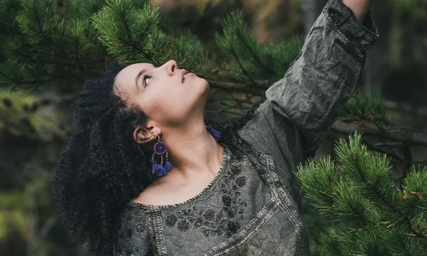 A woman with curly hair reaching upwards among green pine branches, wearing a textured, embroidered blouse and colorful earrings.