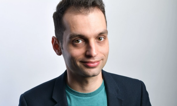 A man with short, dark hair and brown eyes smiles softly, wearing a blue blazer over a teal shirt, set against a light background.