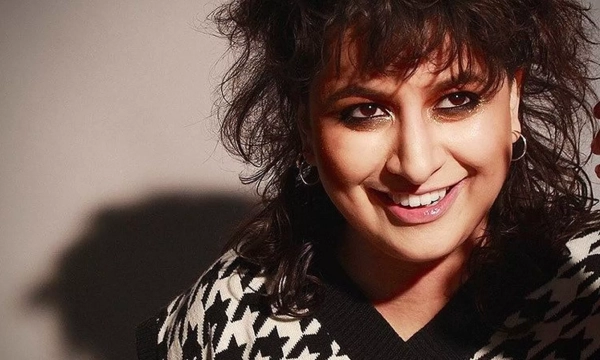 A person with curly hair smiles, wearing a patterned sweater, in a softly lit setting, showcasing a confident and playful expression.