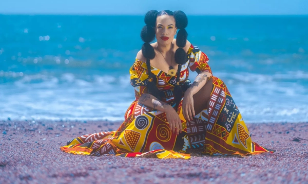 A woman with stylistic puffs in her hair sits on a colorful patterned dress by the beach, with ocean waves in the background.