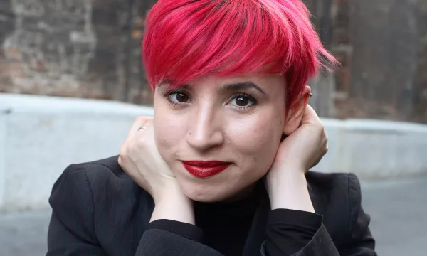 A young person with short pink hair and red lipstick poses with their hands near their face, wearing a black blazer against an urban backdrop.