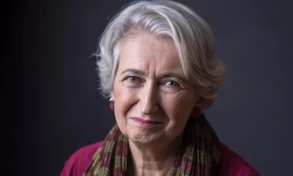 A mature woman with silver hair and a warm smile, wearing a burgundy top and a patterned scarf, against a dark background.