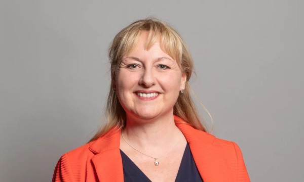 A woman with shoulder-length blonde hair smiles, wearing an orange blazer over a dark top, set against a gray background.