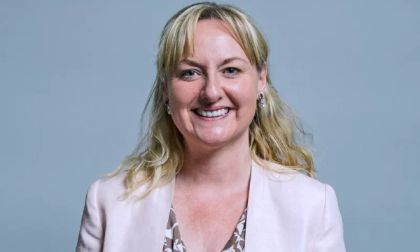 A smiling woman with shoulder-length blonde hair, wearing a light pink blazer over a patterned blouse, poses against a plain grey background.
