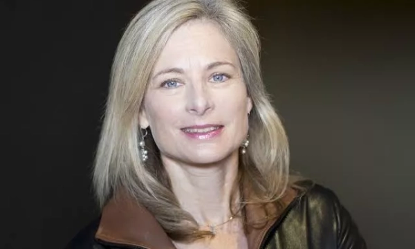 A woman with long blonde hair wearing a black leather jacket and earrings, smiling against a dark background.