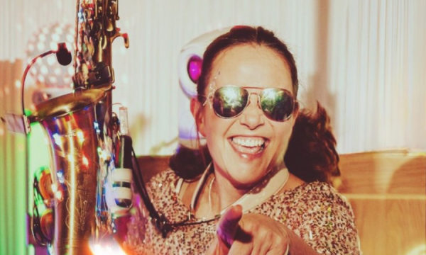 A lively musician wearing sunglasses holds a saxophone and smiles while pointing at the camera, surrounded by colorful party lights.