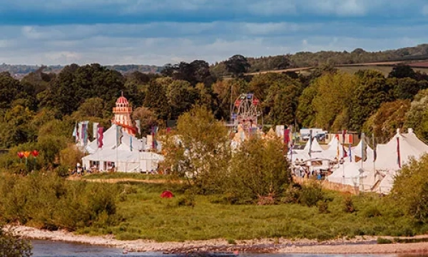 A picturesque view of a festival by a river, featuring white tents, colorful flags, and greenery in the backdrop against a cloudy sky.