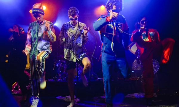 A group of five performers dressed in colorful, eclectic outfits dancing together on stage, with vibrant stage lighting enhancing the energetic atmosphere.