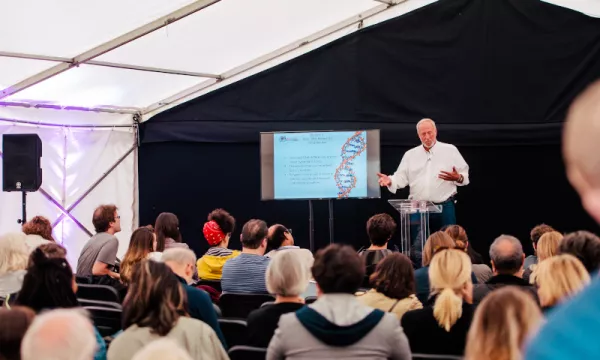 A speaker presents in a tent to an audience, with a screen displaying a DNA-related graphic behind him.