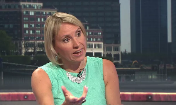A woman in a green dress gestures while speaking during a discussion, with a city skyline visible in the background.