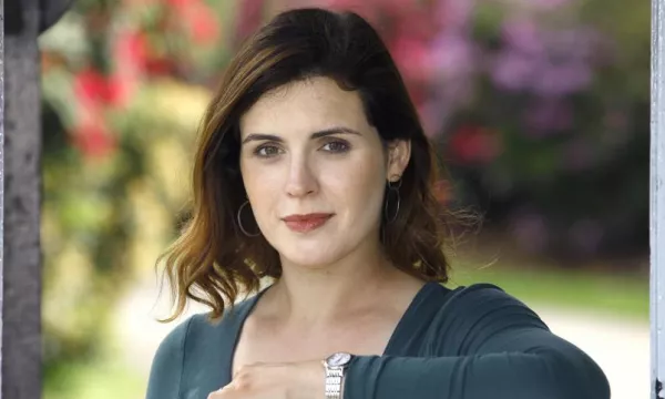A woman with shoulder-length brown hair poses in a garden setting, wearing a green top and silver watch, with colorful flowers blurred in the background.