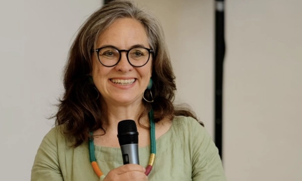 A smiling woman with glasses and long, wavy hair holds a microphone, wearing a green top and colorful beaded necklace.