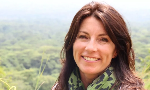 A smiling woman with long brown hair and a green patterned scarf, standing against a lush green landscape.