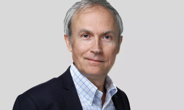 A middle-aged man with gray hair wearing a dark blazer over a light checkered shirt, smiling against a plain gray background.