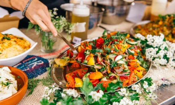 A vibrant platter of mixed vegetables and herbs is being served, surrounded by bowls of food and decorative flowers on a rustic table.