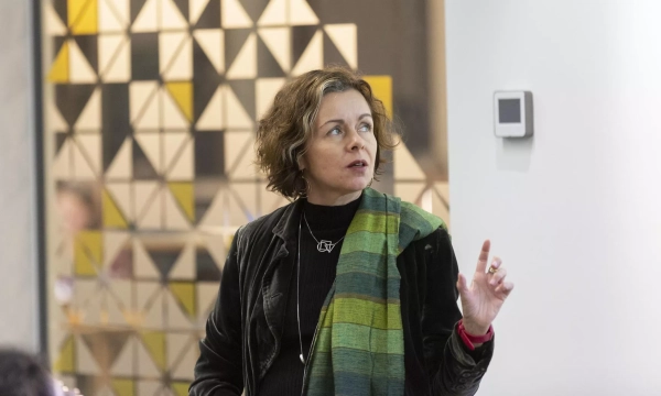 A woman with curly hair gestures while speaking, wearing a black jacket and a colorful scarf, with geometric patterned decor in the background.