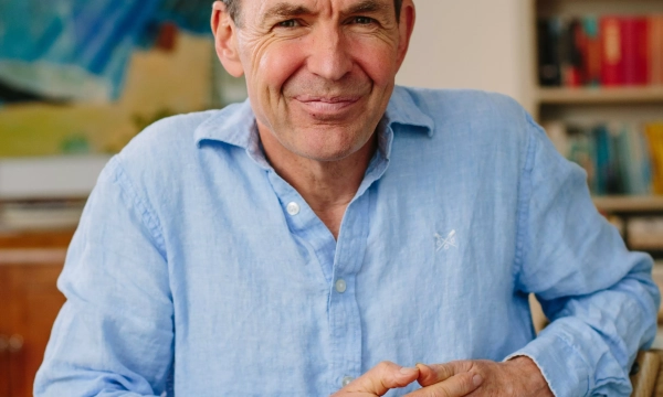 A smiling man in a light blue shirt sits with his hands clasped at a table, in a bright room decorated with books and art.