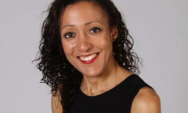 A woman with curly hair smiles warmly at the camera, wearing a black sleeveless top against a neutral background.
