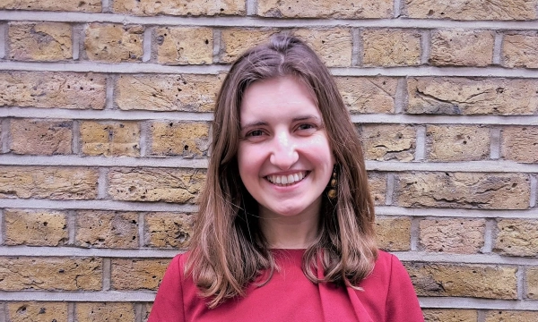 A smiling woman with medium-length brown hair stands in front of a textured brick wall, wearing a red top.