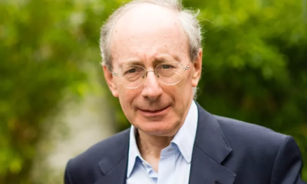 A man with gray hair and glasses, wearing a dark blazer over a light shirt, stands outdoors with greenery in the background.