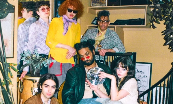 A diverse group of six individuals poses in a stylish, retro-inspired setting, showcasing vibrant fashion and unique hairstyles with musical instruments present.