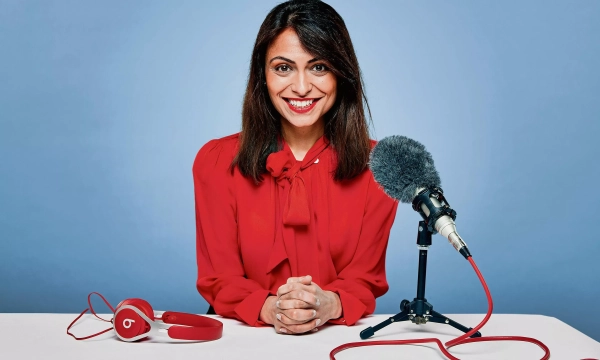 A confident woman in a red blouse sits at a table with a microphone and headphones, smiling against a light blue background.