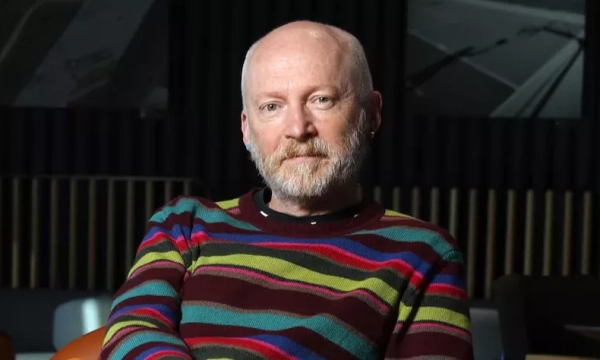 A man with a beard wearing a colorful striped sweater sits in a modern indoor setting, reflecting a thoughtful demeanor.