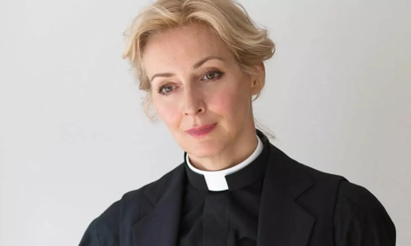 A woman dressed in a clerical collar and black suit, portraying a modern clergyperson, with a thoughtful expression against a plain background.