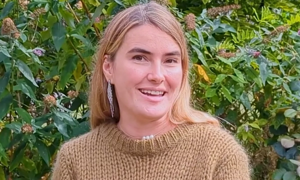 A smiling woman wearing a cozy sweater stands outdoors amidst lush green foliage and flowering plants.