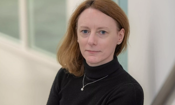 A woman with shoulder-length reddish-brown hair wearing a black turtleneck sweater sits in a well-lit environment, facing the camera with a neutral expression.