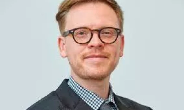 A professional-looking man with short, light brown hair and glasses, wearing a black suit with a checked shirt and a tie, smiles confidently.