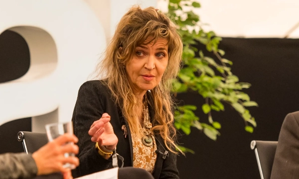 A woman with long, wavy hair speaks passionately at a conference or panel discussion, gesturing with her hand, surrounded by greenery.