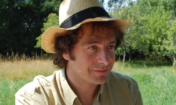 A man with curly hair wearing a straw hat and a light-colored shirt is sitting in a sunny outdoor setting surrounded by greenery.