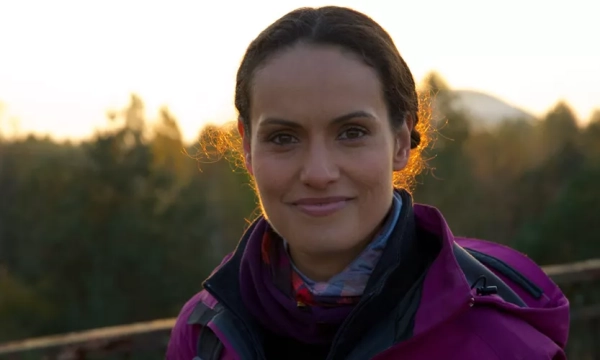 A smiling woman with long, dark hair stands against a sunset backdrop, wearing a purple outdoor jacket and a multi-colored scarf, surrounded by trees.