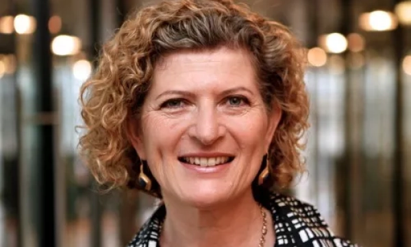 A smiling woman with curly hair, wearing a patterned black and white shirt, stands in a modern setting with blurred lights in the background.