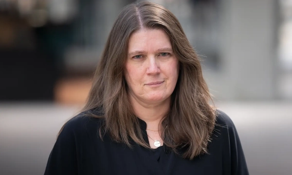 A woman with long brown hair wearing a black blouse stands outdoors, looking directly at the camera with a serious expression.